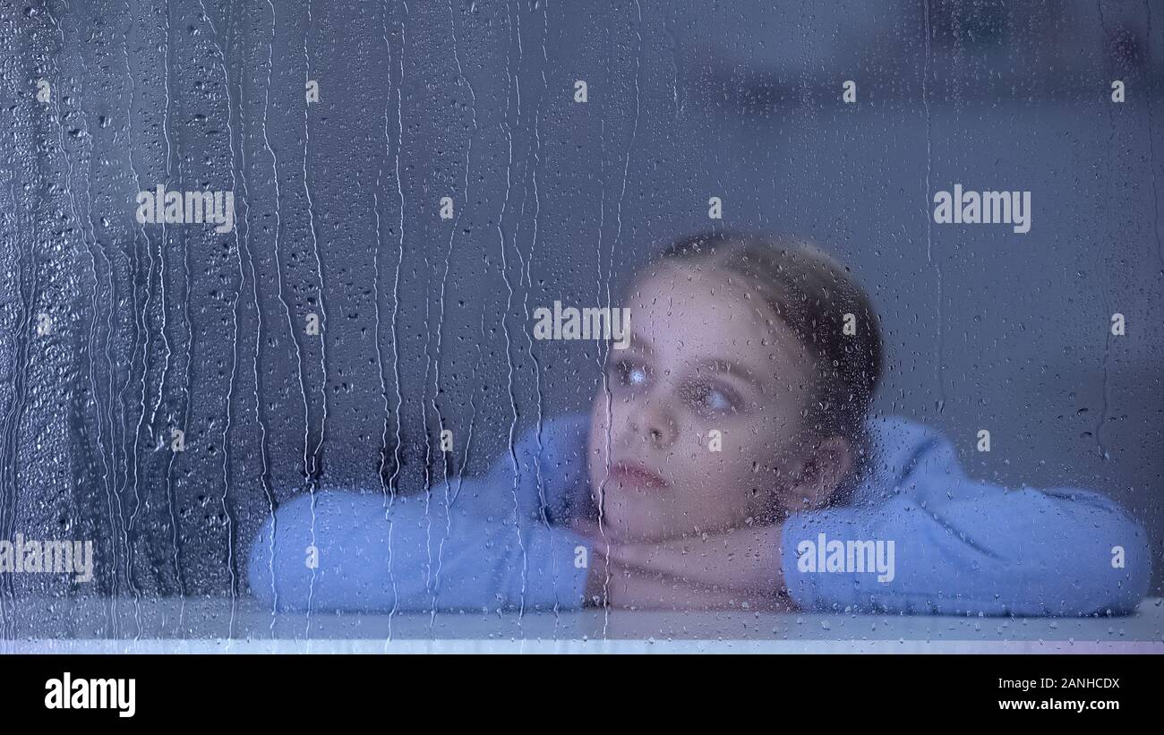 Lonely girl cerca attraverso la finestra piovosa mancanza di amore parentale e di cura, noia Foto Stock
