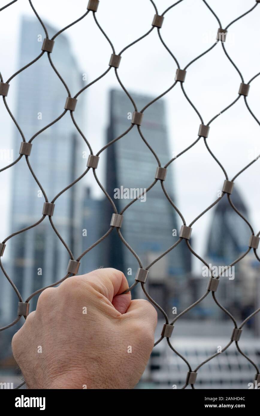 Stretta di mano d'uomo afferrato wire mesh recinzione contro la città di Londra edifici in background come urban inquietudine e incertezza concetto. Foto Stock