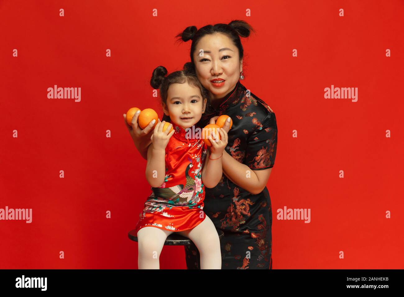 Abbraccia, sorridente, tenendo mandarini. Felice Anno Nuovo Cinese 2020. Asian madre e figlia ritratto su sfondo rosso in abiti tradizionali. Celebrazione, emozioni umane, vacanze. Copyspace. Foto Stock