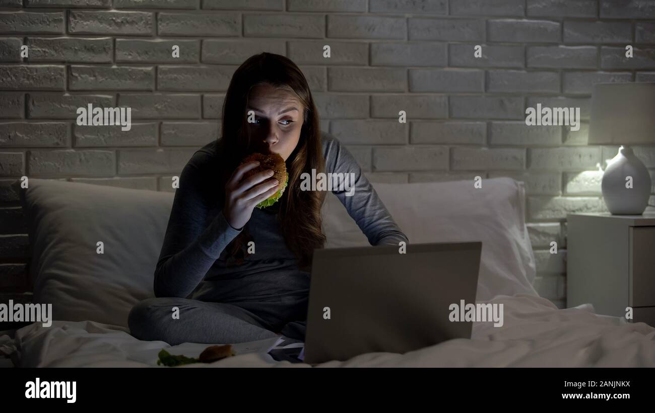 Giovane donna mangiare cheeseburger e guardare film interessante su laptop, tempo libero Foto Stock