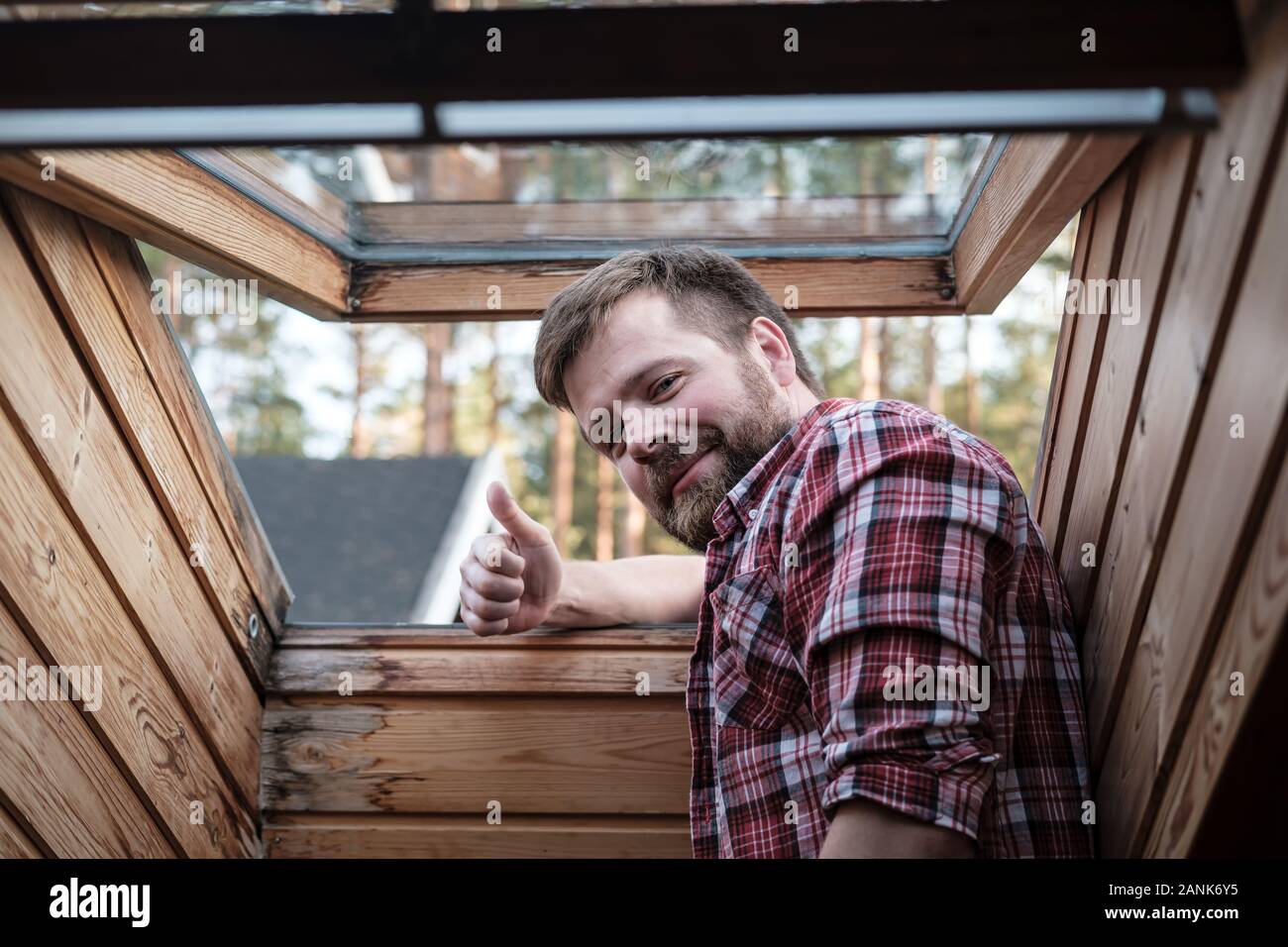 Uomo Barbuto sorge nelle vicinanze di un tetto aperto nella finestra mansarda e fa un gesto di approvazione, pollice in alto, sorridente e guardando la telecamera. Foto Stock