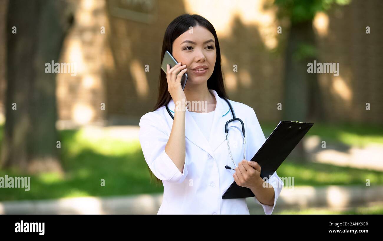 Piuttosto medico parlando con paziente sullo smartphone in ospedale park, 24h Foto Stock
