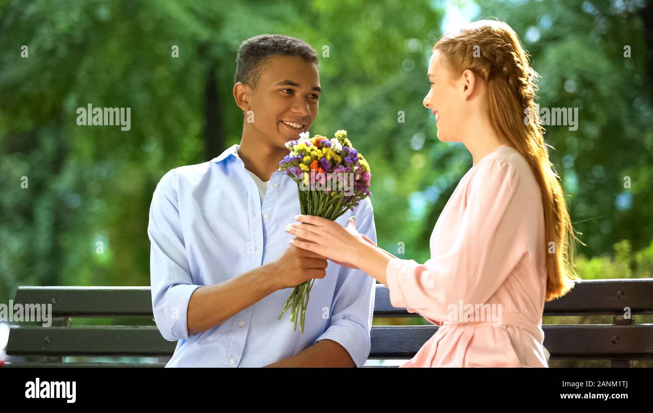 Sorridente mixed-gara teen boy presentando cara ragazza fiori, prima data in posizione di parcheggio Foto Stock