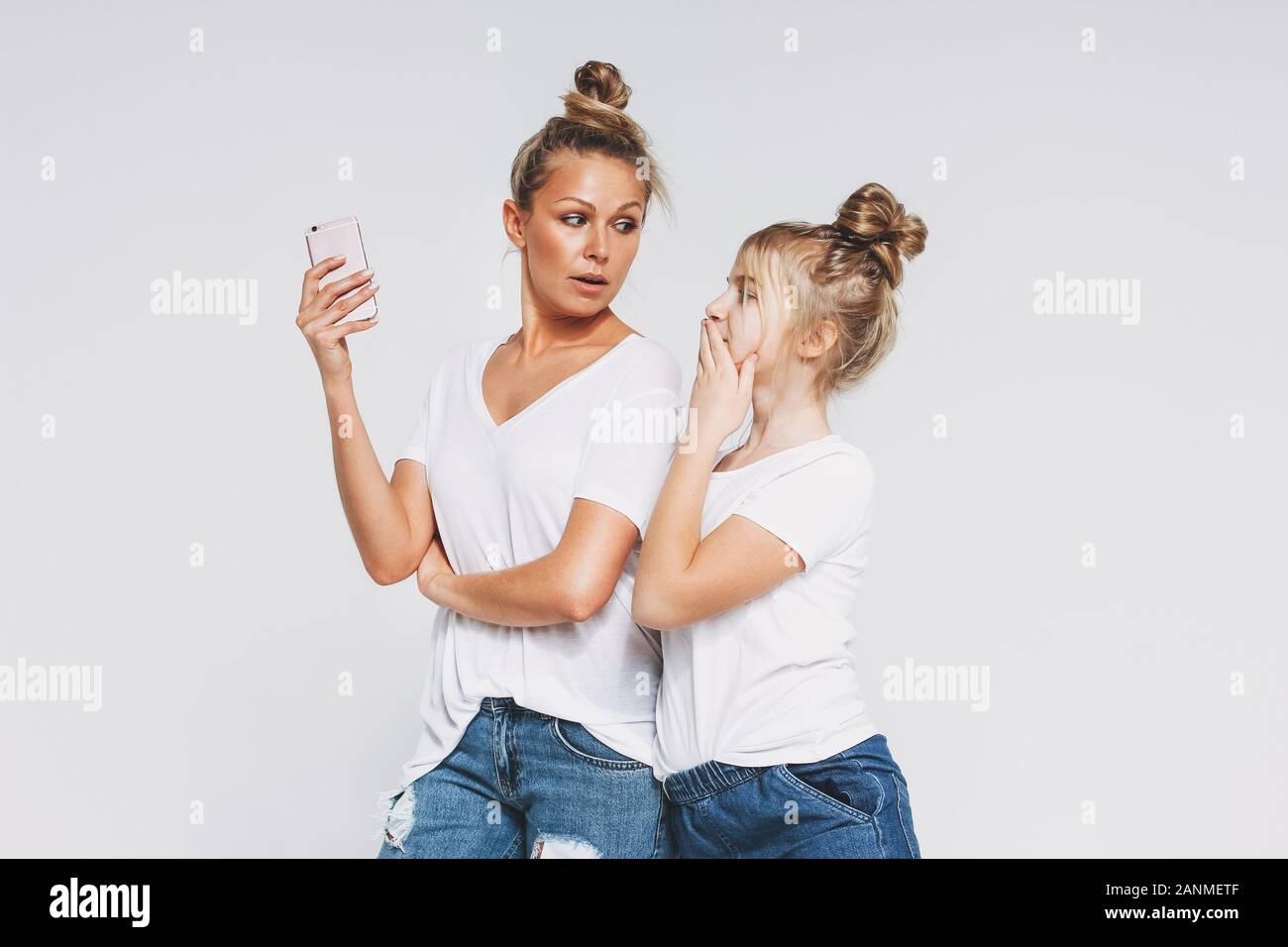 Blonde sorpreso mamma e figlia in bianco t-shirt e jeans utilizzando i telefoni cellulari gadget concetto isolato su sfondo bianco Foto Stock