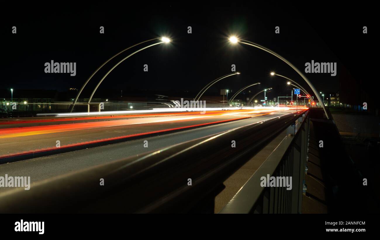 Rosso e bianco auto sentieri dei fari di notte sul ponte, racchiuso dai lampioni curvi; le linee diagonali di fornire velocità e profondità Foto Stock