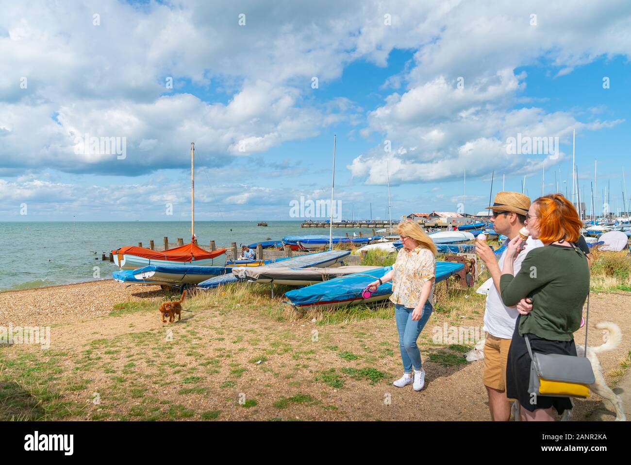 Whitstable Inghilterra - Il 18 agosto 2019; la gente a mangiare il gelato e con il cane sui conduttori in piedi sul lungomare dove piccole barche e yacht sono sulla spiaggia. Foto Stock