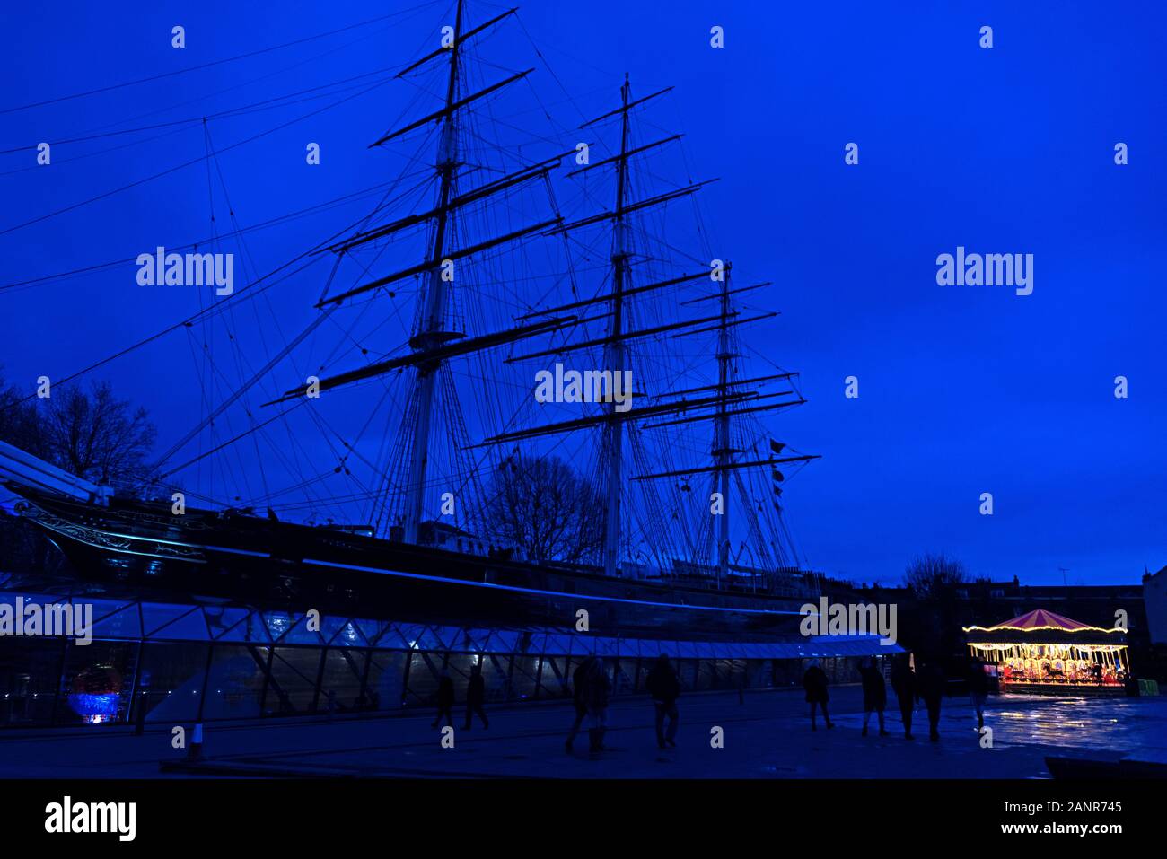 Luci di una giostra incandescente in inverno il crepuscolo scompaiono di fronte alla silhouette del nearcy Cutty Sark, Greenwich, Inghilterra. Foto Stock