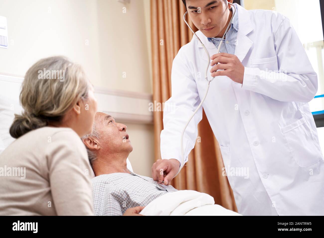 Asian anziani paziente di sesso maschile che giace nel letto che viene esaminato dal giovane medico in ospedale Foto Stock