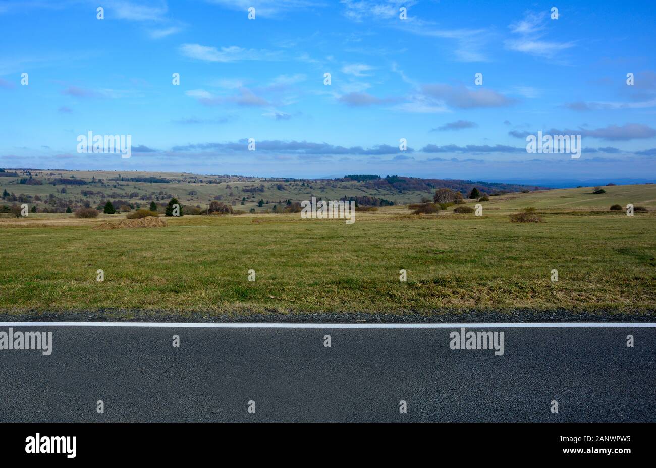 Paesaggio con parte di una strada nella parte inferiore dell'immagine, con un grande prato con alberi e cielo blu nel Rhön, Baviera, Germania Foto Stock