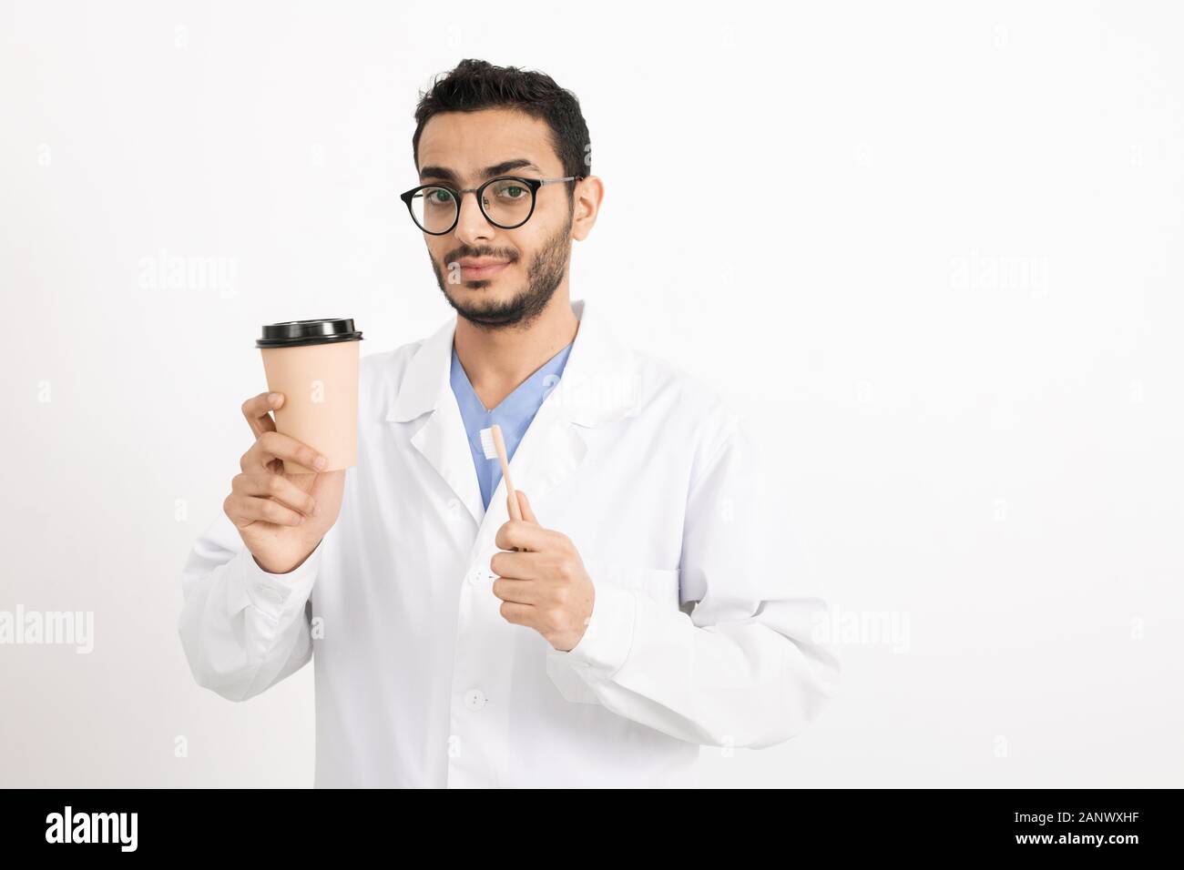 Giovani dentista in camice bianco e occhiali tenendo un bicchiere di caffè e spazzolino da denti Foto Stock