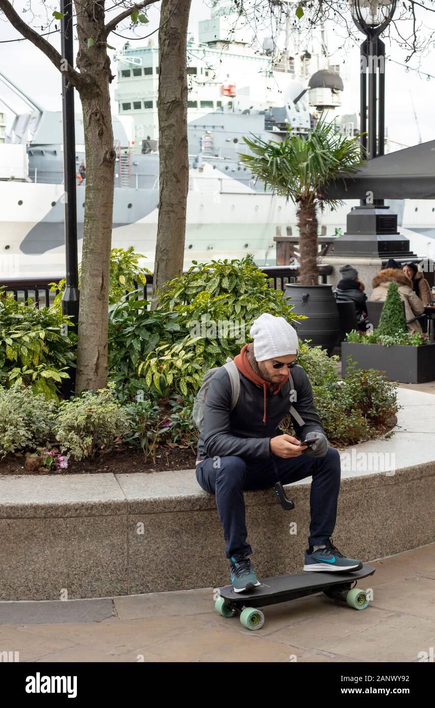 Giovane uomo con skateboard elettrico potenziato Mini X e controllo bluetooth intorno al polso seduto e fare una pausa nella South Bank Londra, Regno Unito Foto Stock