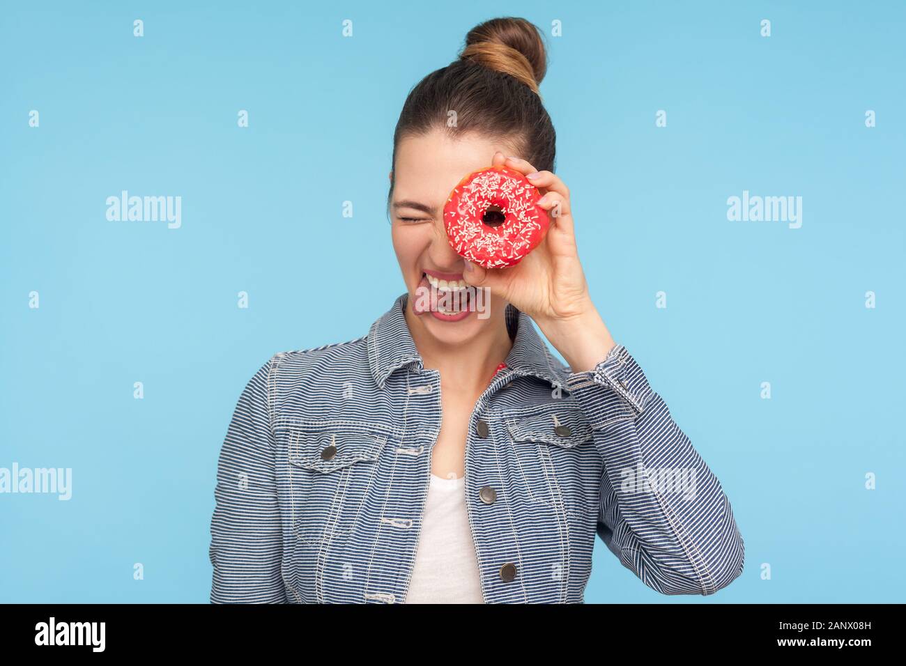 Primo piano di carefree divertito ragazza con i capelli bun spiata attraverso la ciambella e spuntavano lingua, rendendo faccia avendo divertimento con snack, cercando di ciambella. Foto Stock