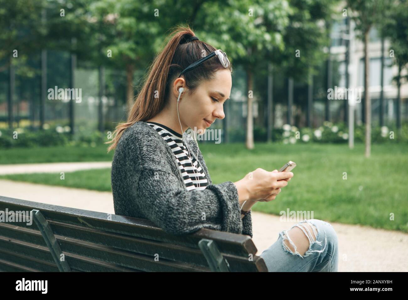 Una giovane bella ragazza si siede su un banco in un parco della città e per la riproduzione di musica o di un podcast o apprende da lezioni audio. Foto Stock