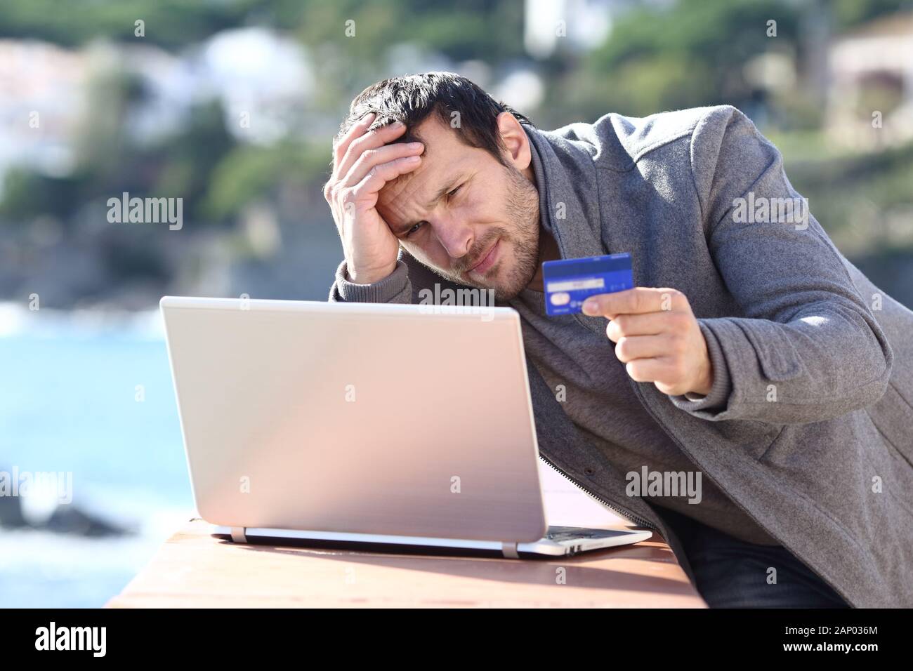 Preoccupato uomo in inverno acquisti on line con carta di credito e il computer portatile in un balcone in una città costiera Foto Stock
