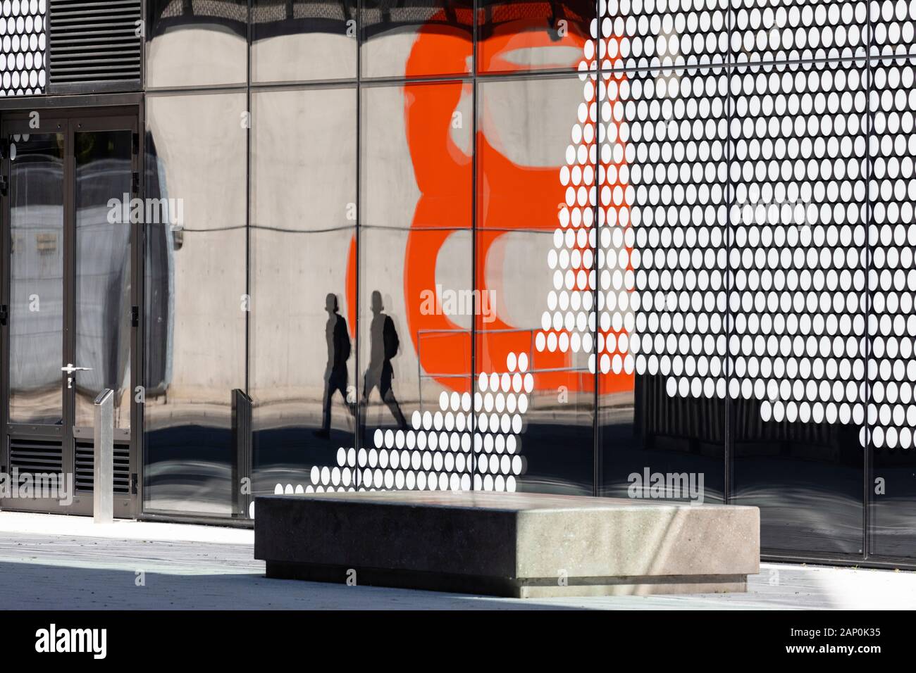 Una solitaria figura si riflette nella grafica facciata di vetro di un edificio moderno in qui est campus presso la Queen Elizabeth Olympic Park. Foto Stock