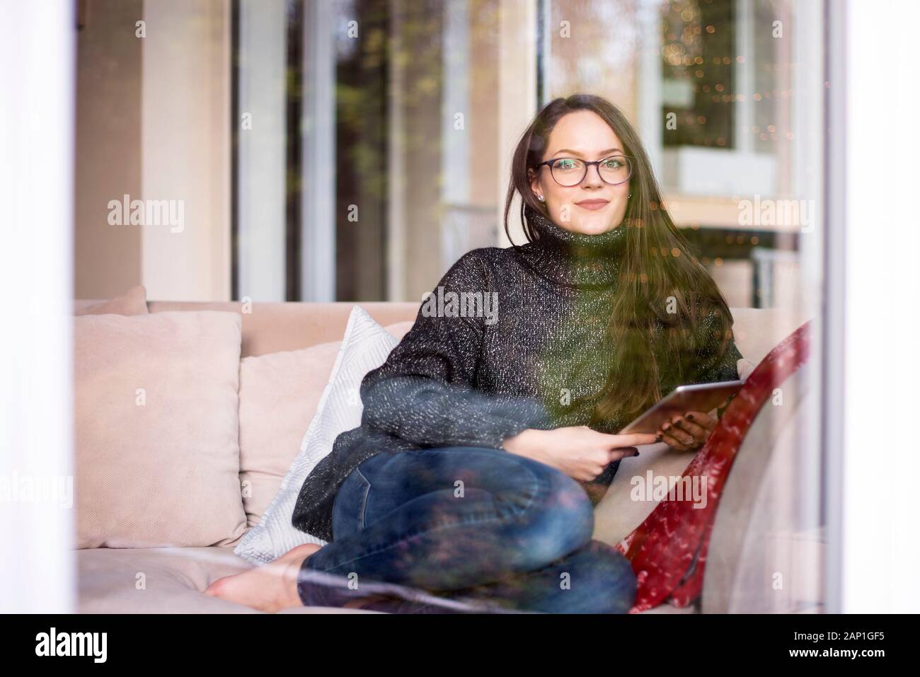 Colpo di donna sorridente dietro la finestra di vetro navigando in rete sulla tavoletta digitale durante i momenti di relax in poltrona a casa. Foto Stock