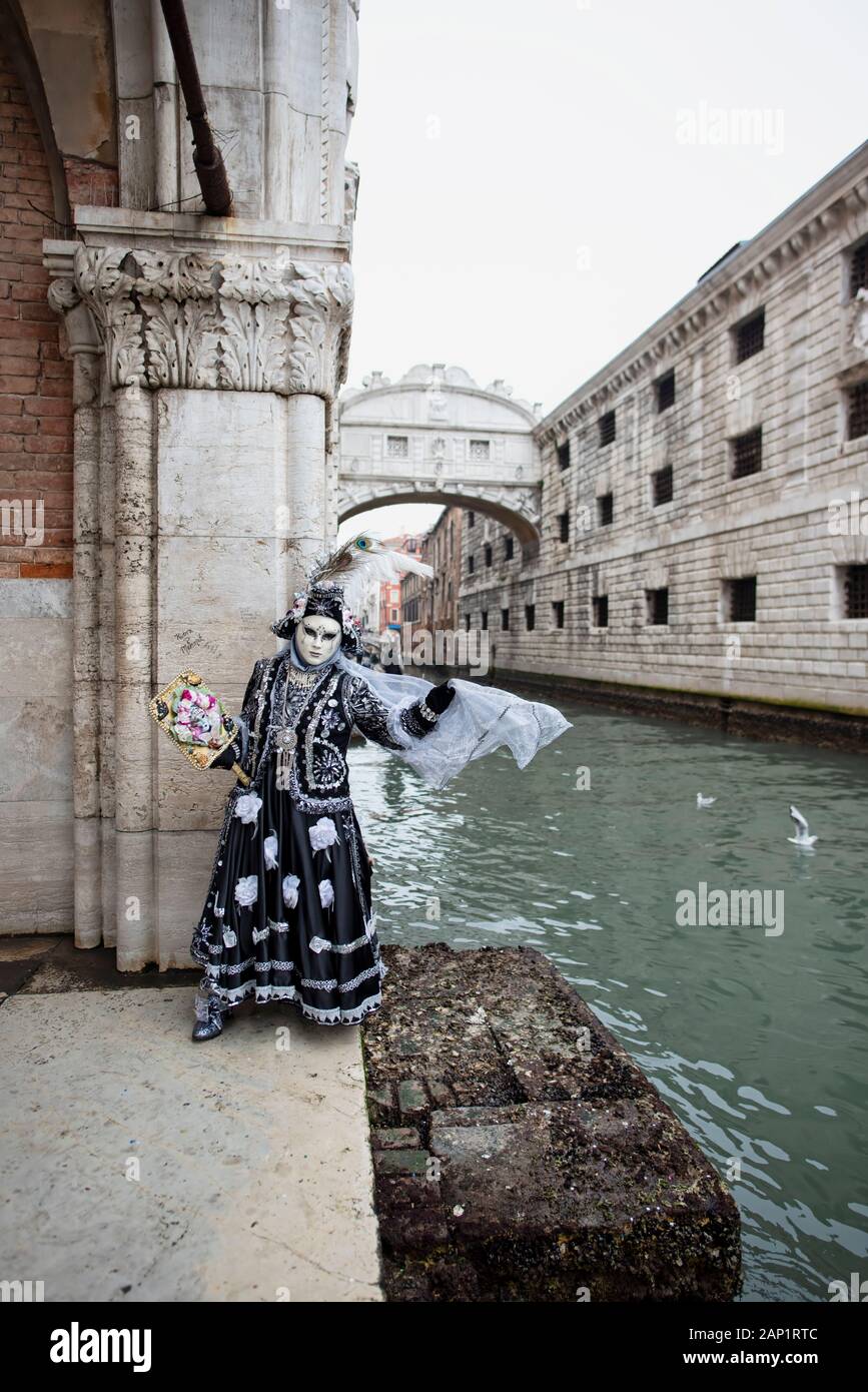 Una persona in maschera di carnevale appoggiata su un muro di un edificio con il ponte degli avvistamenti sullo sfondo Venezia Italia Foto Stock