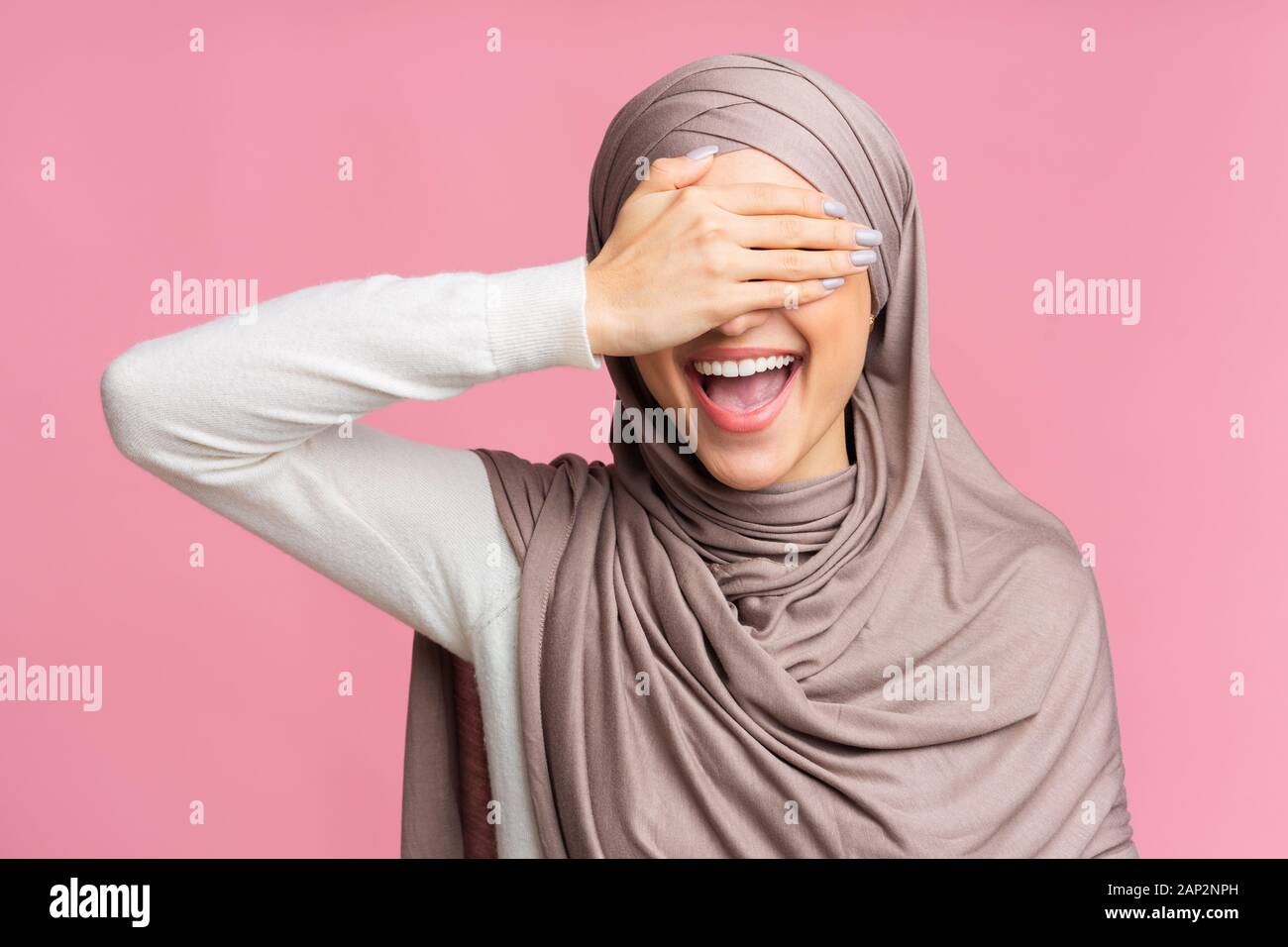 Felice ragazzina musulmana in Hijab che copre gli occhi con la mano e sorridente, divertirsi su sfondo rosa, spazio libero Foto Stock