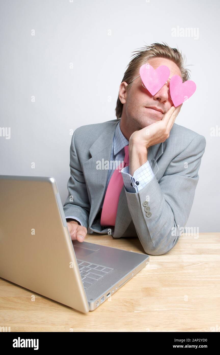 Imprenditore lavorando sul suo computer portatile fantasticando con cuori rosa svolazzanti oltre i suoi occhi Foto Stock