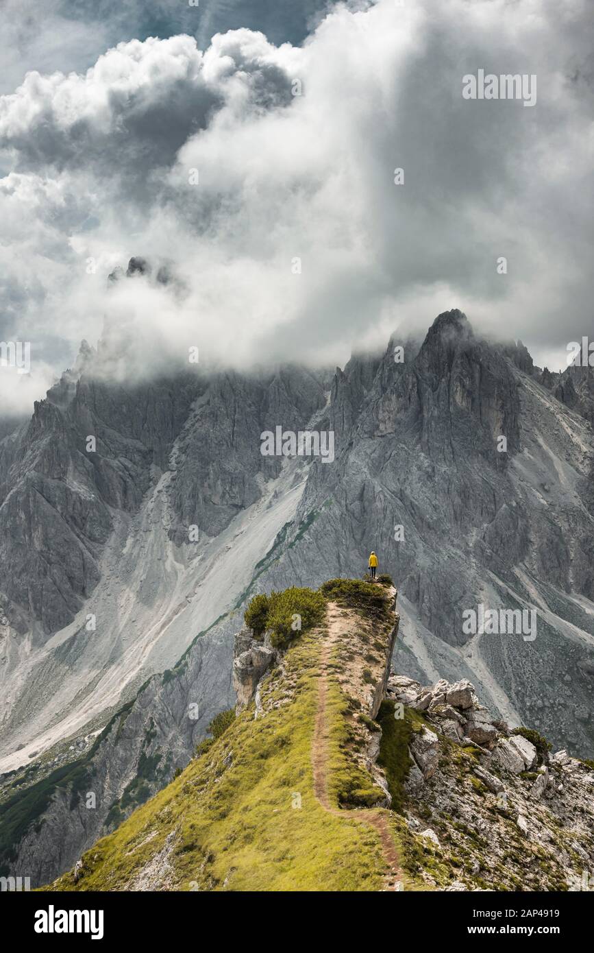 Donna in giacca gialla in piedi su un crinale, dietro il suo volto di roccia, nuvole drammatiche, Cimon la Croda liscia e il gruppo Cadini, Auronzo di Cadore Foto Stock