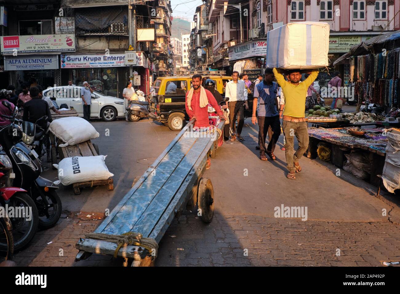 Un portiere spinge il suo carrello attraverso la trafficata area bazar di Bhendi a Mumbai, India, un altro che trasporta merci sulla sua testa Foto Stock