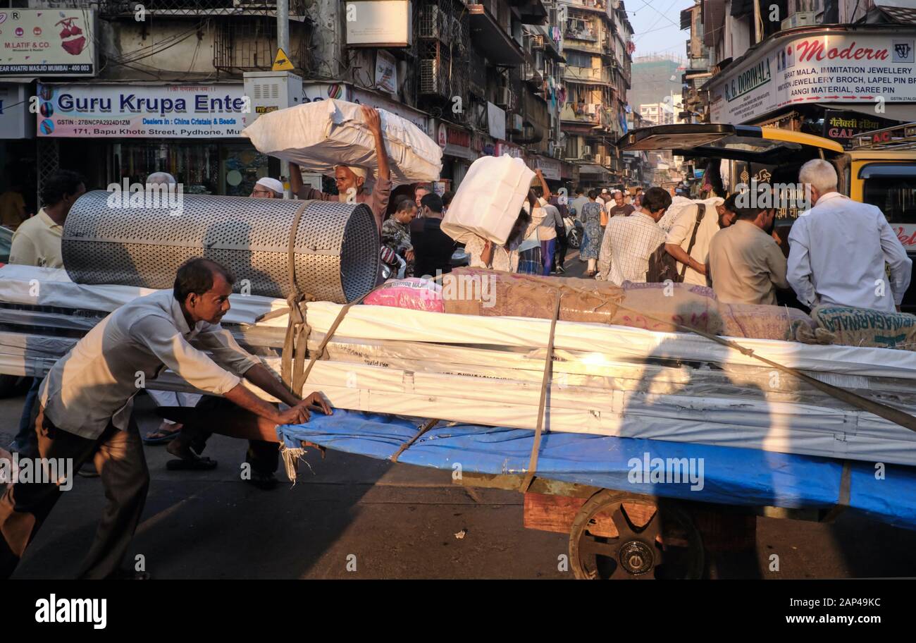 Un portiere spinge il suo carrello attraverso la trafficata area bazar di Bhendi a Mumbai, India, altri che trasportano merci sulla testa Foto Stock