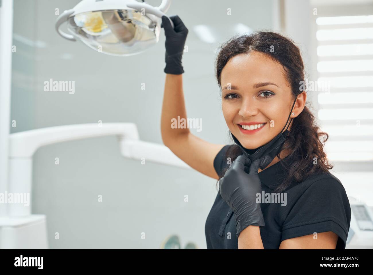 Ritratto di bella dentista femminile in uniforme medica nera sorridente e guardando la fotocamera con sfondo sfocato del cabinet dentale. Felice donna sicura al posto di lavoro. Foto Stock