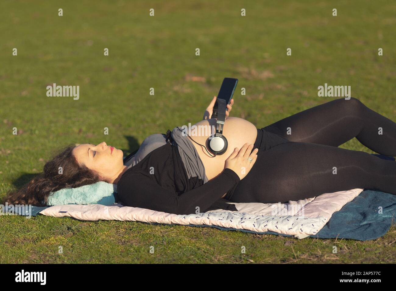 Donna incinta rilassante in un campo di erba con cuffie oltre il suo ventre per dare un bambino nel grembo di una possibilità di ascoltare la musica Foto Stock