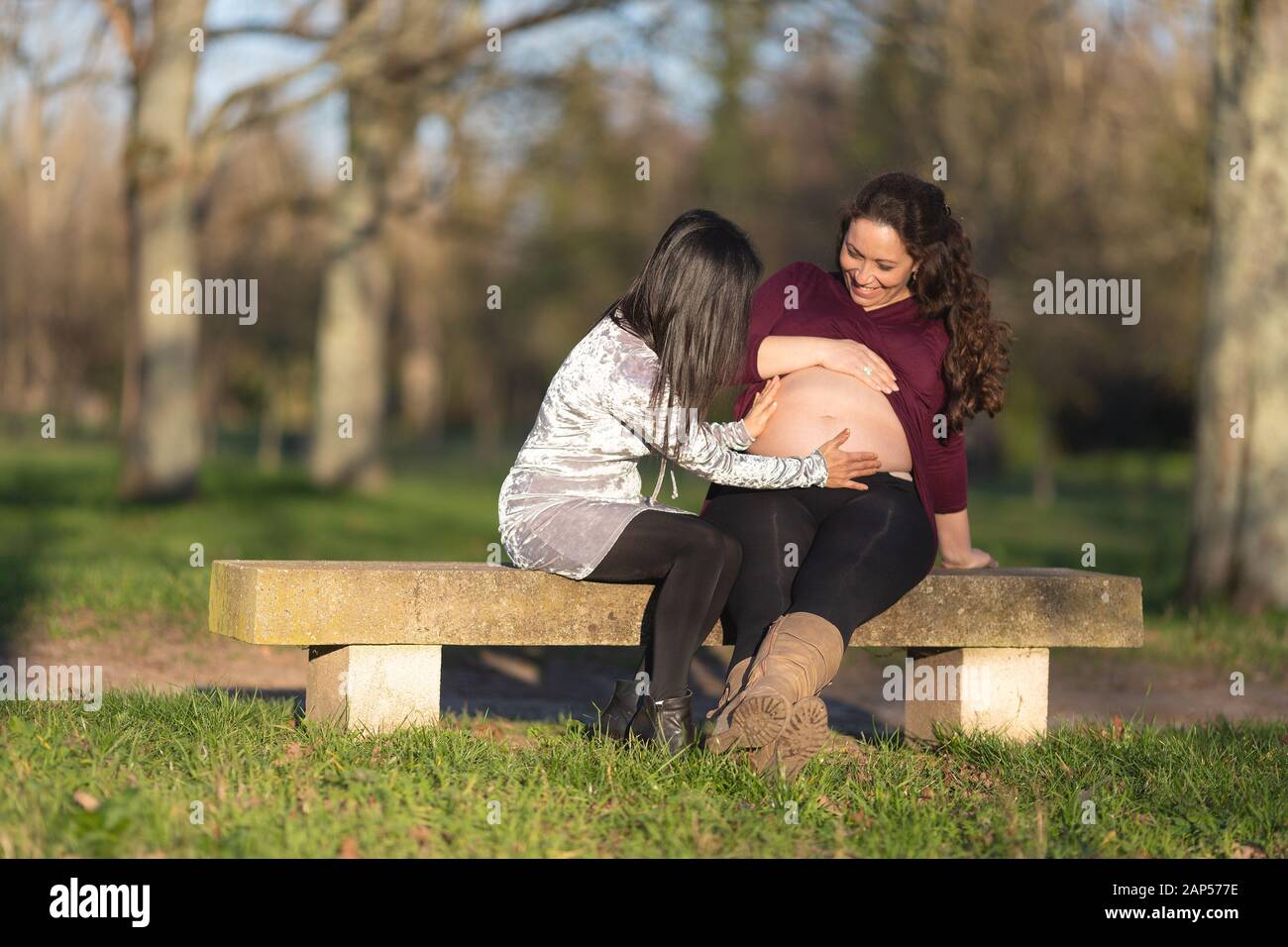 Giovane donna incinta mostra il suo baby bump o gonfiore addominale di un amico su una panchina nel parco con una felice amorevole sorriso Foto Stock