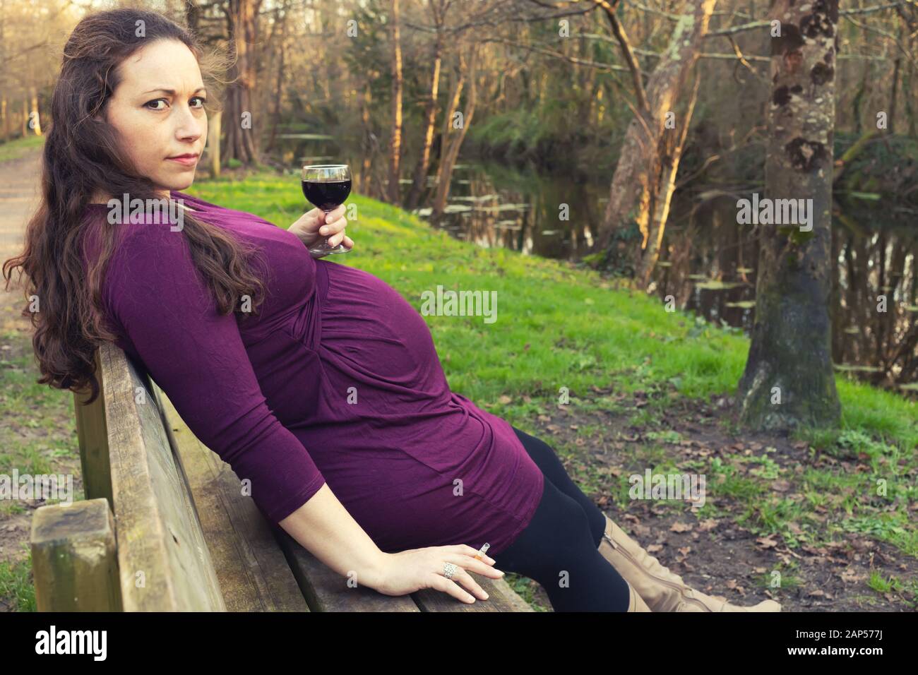 Donna incinta con alcool e sigarette nelle sue mani, seduti su una panchina nel parco e guardando la telecamera con un interrogatorio guardare . Lato verticale con Foto Stock