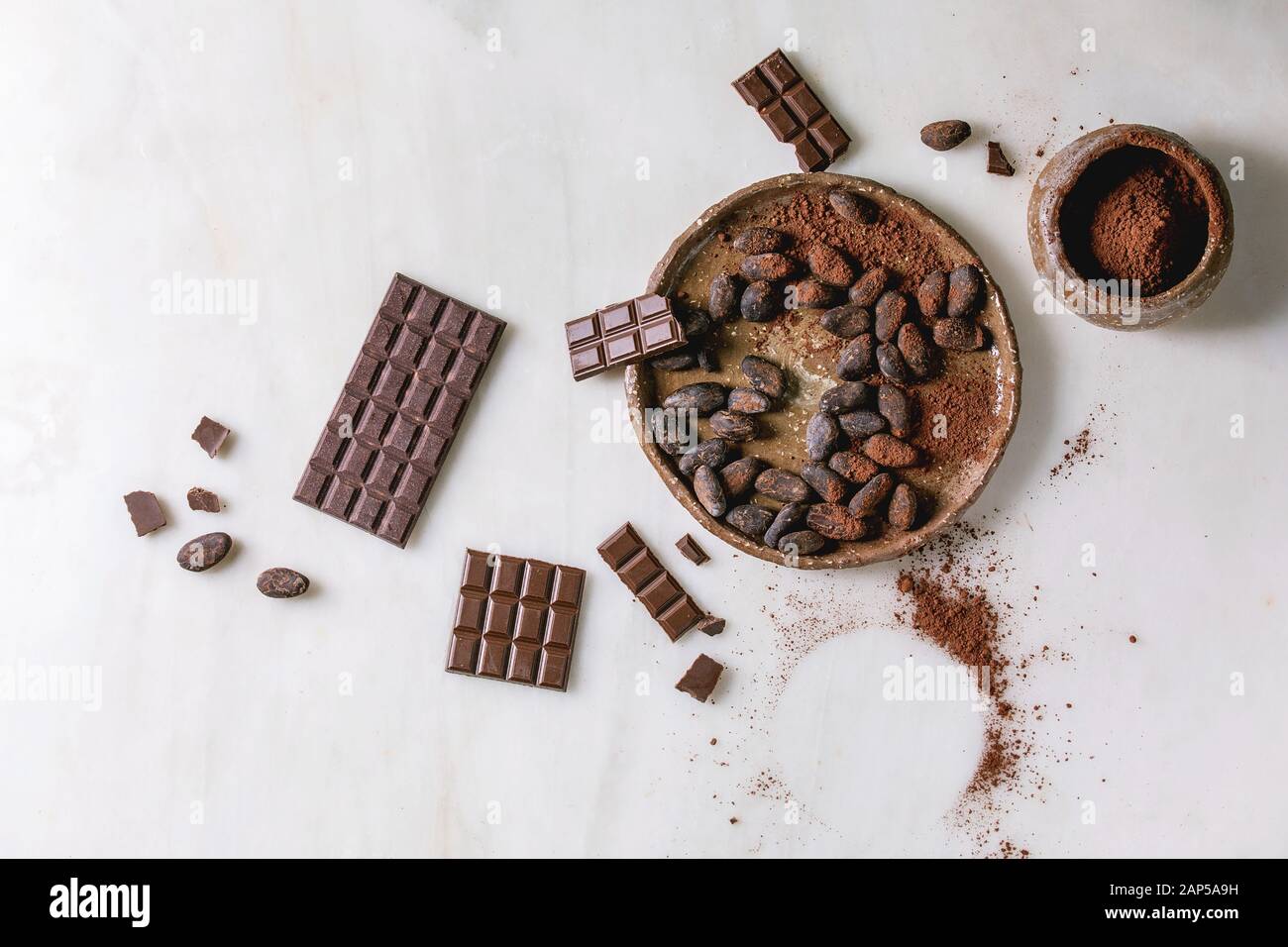 Cioccolato fondente e al latte intero bar e tritate con le fave di cacao, cacao in polvere nella ciotola in ceramica bianca su sfondo marmo. Laici piana, spazio Foto Stock