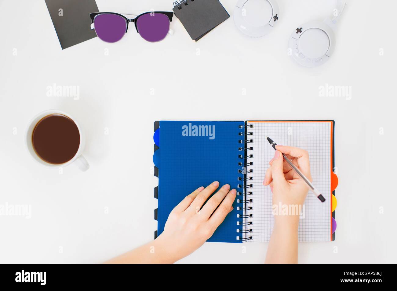 Flatlay con le mani femminili che scrivono in un notebook. Spazio di lavoro minimo per l'ufficio. Sfondo bianco e tazza di caffè nero accanto. Foto Stock