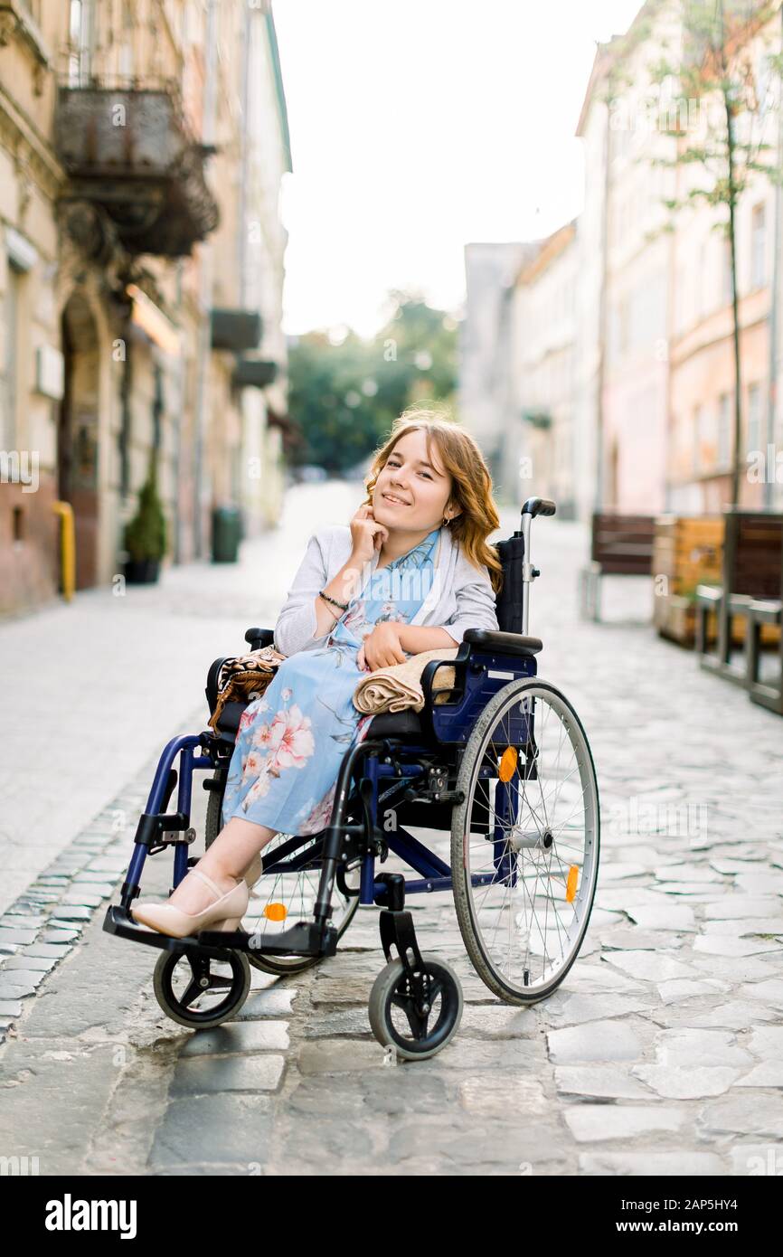 bella ragazza bionda per disabili in abito blu seduta su sedia a rotelle, all'aperto in città. Foto Stock