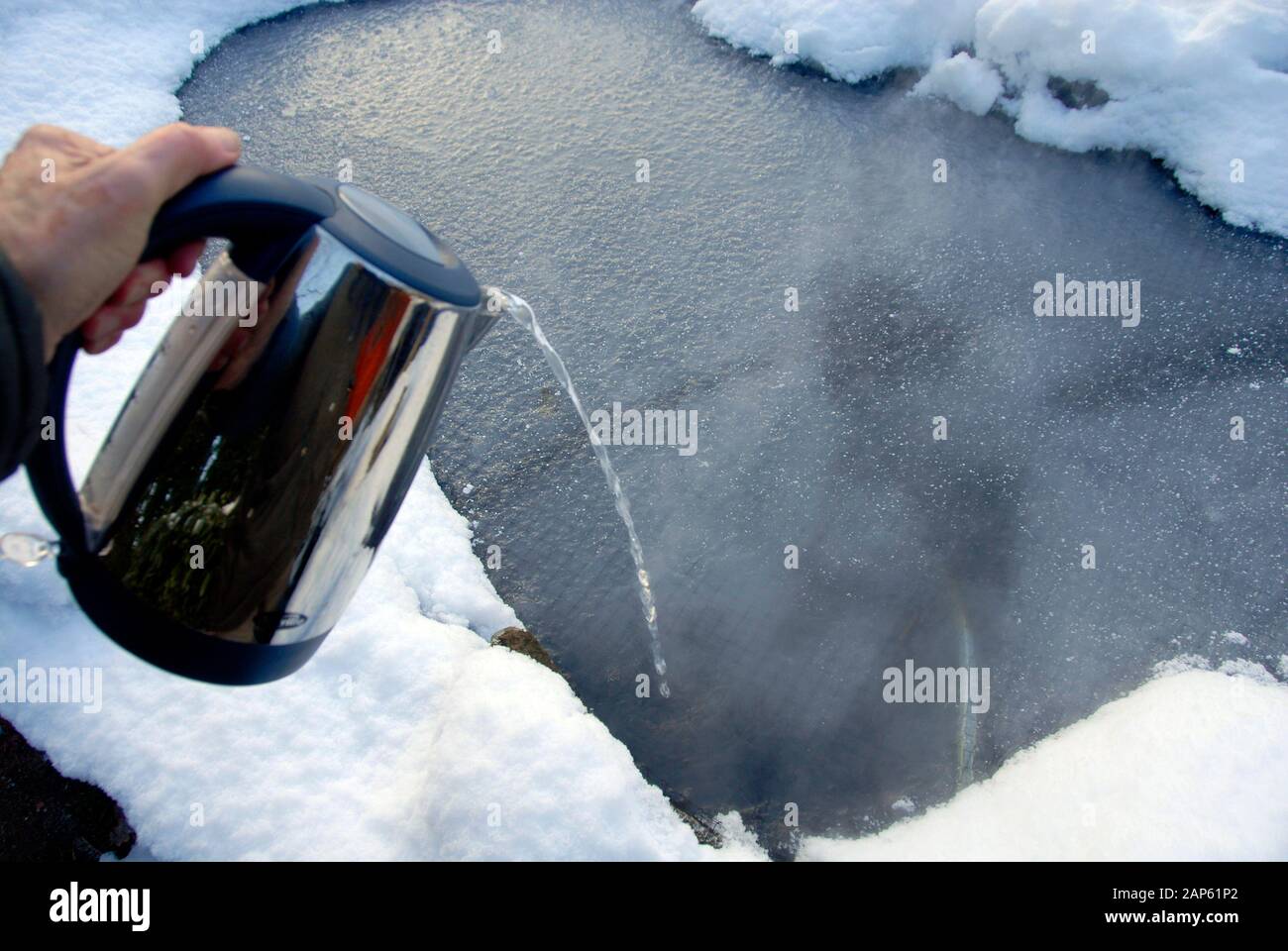 Versare acqua molto calda da un bollitore elettrico per sciogliere il ghiaccio formatosi sul laghetto del giardino domestico. Foto Stock