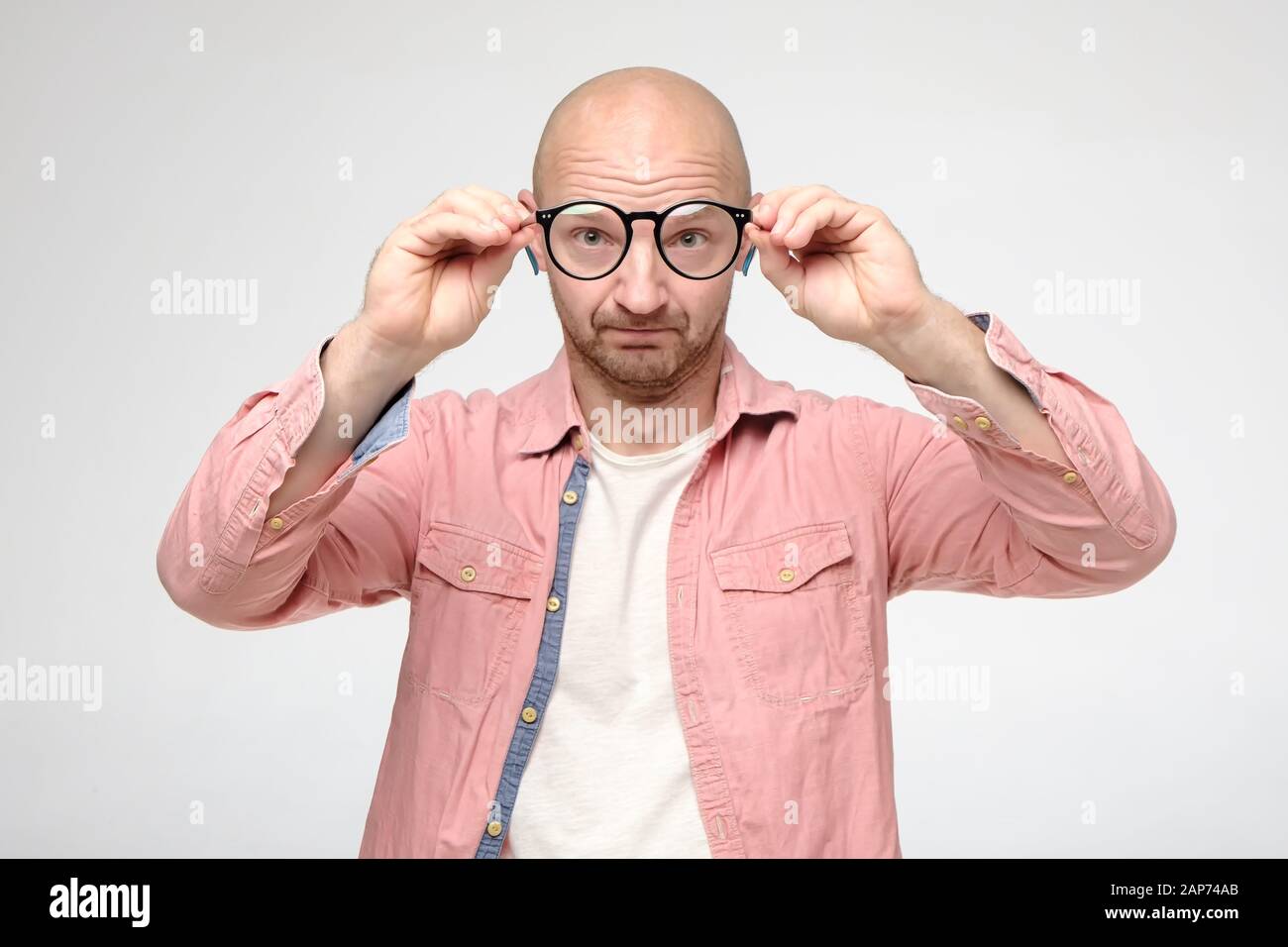 Bald, unshaven uomo ha tolto gli occhiali, mantenendoli ad una distanza, esamina la pulizia delle lenti e sembra divertente. Foto Stock