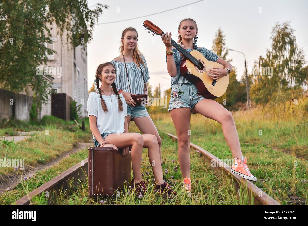 Tre ragazze adolescenti stand sui binari del treno con una chitarra, una vecchia valigia marrone e una fotocamera. Foto Stock