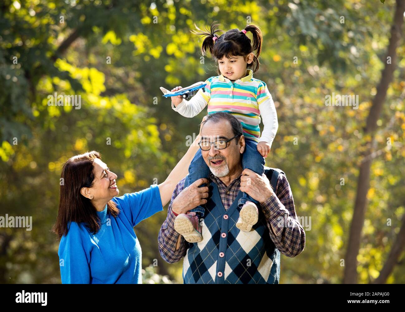Nonna con ragazza che tiene l'aeroplano giocattolo seduto sulla spalla del nonno al parco Foto Stock