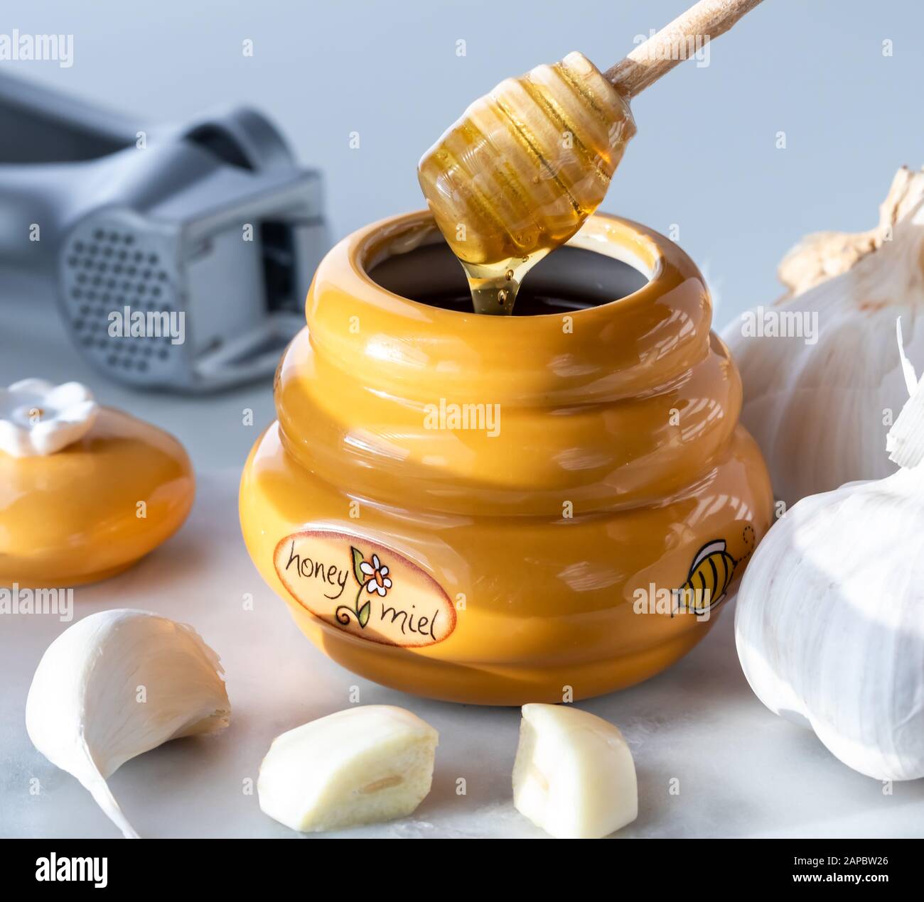 Una vista ravvicinata di un piatto di miele con un cucchiaia che gocciola miele e circondato da aglio e un pressa di aglio. Foto Stock