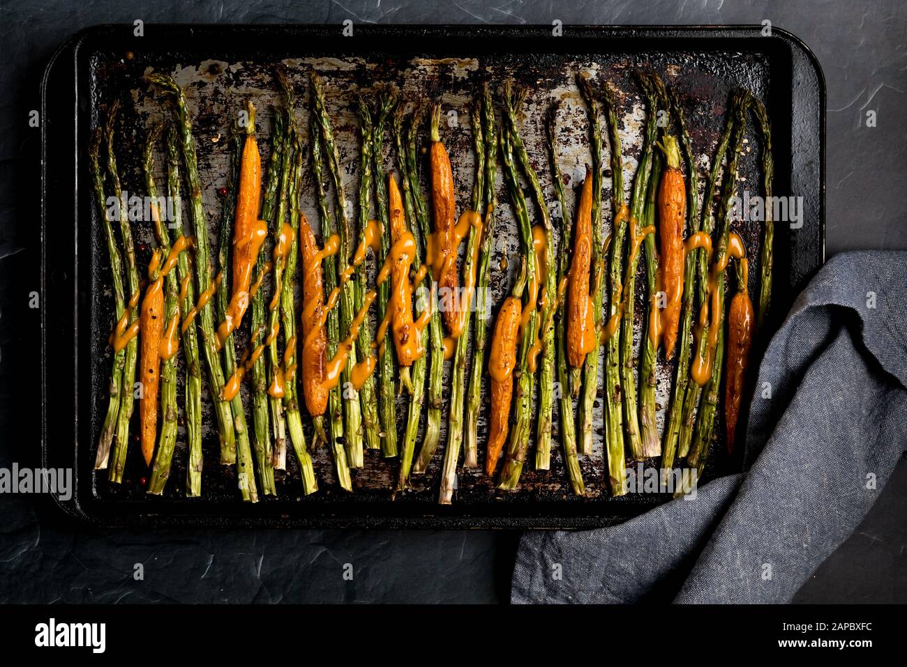 Una vista dall'alto verso il basso di un foglio di cottura con asparagi arrostiti al forno e carote al bambino condite con salsa. Foto Stock