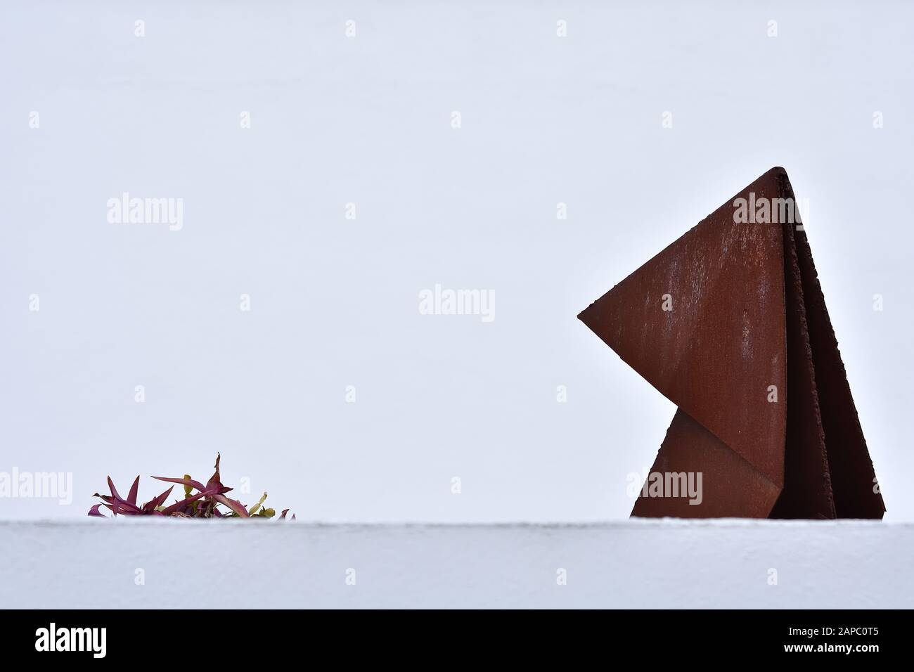 Figura decorativa metallica che si trova accanto ad una piccola pianta dietro un muro con uno sfondo completamente bianco, dando un'impressione minimalista Foto Stock