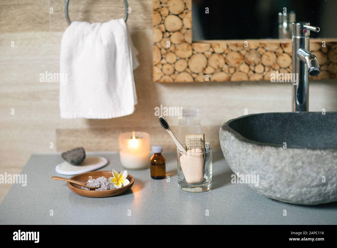 Accessori da bagno naturali: Spazzolino da denti in bambù, prodotto per la rimozione in un contenitore di vetro, candela di cera di cocco, sale da bagno organico, pomice vulcanico e asciugamano di cotone in piedi vicino al lavello di pietra nera. Foto Stock