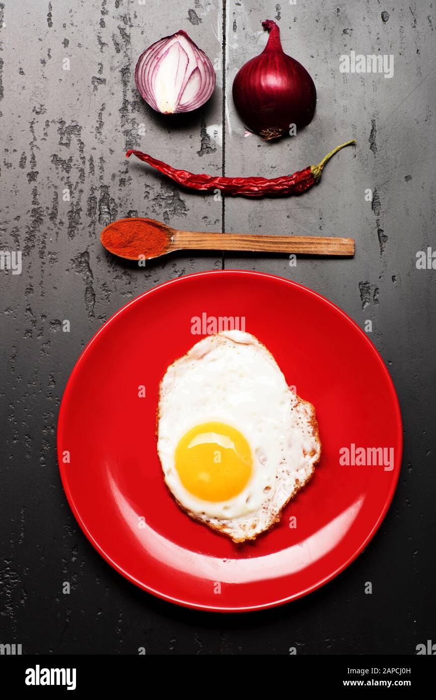 Cibo collocato su piastra rossa su sfondo nero, vista dall'alto. Colazione e cibo art concetto. Frittata messa al centro della piastra. Piatto con uovo vicino a cucchiaio con paprika, peperoncino rosso e cipolle che fanno faccia Foto Stock