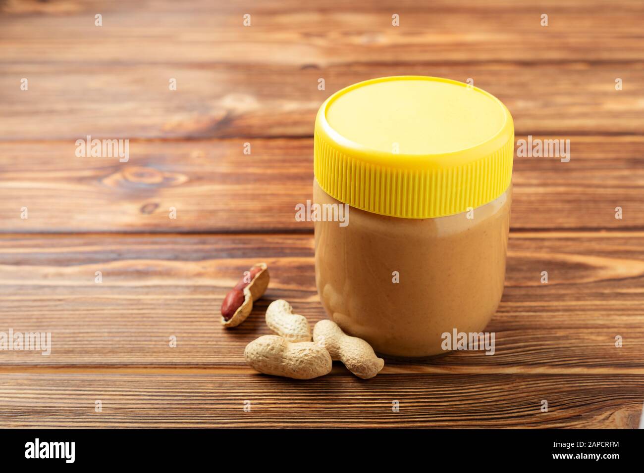Pasta di arachidi cremosa in vaso di vetro con tappo giallo e arachidi nella buccia sparsi sul tavolo di legno marrone con spazio copia per la colazione di cottura Foto Stock