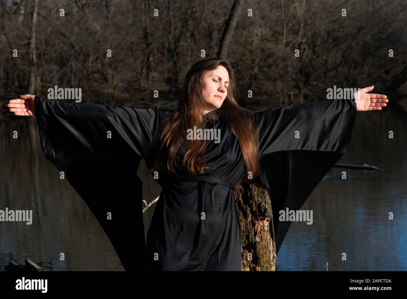 Donna con capelli lunghi scuri in abiti neri di fronte al lago. Magia e Witchcraft. Torna al concetto di natura. Foto Stock