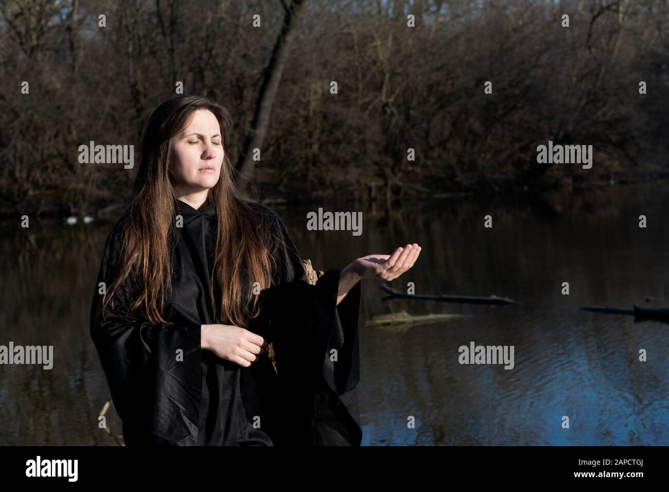 Donna con capelli lunghi scuri in abiti neri di fronte al lago. Magia e Witchcraft. Torna al concetto di natura. Foto Stock