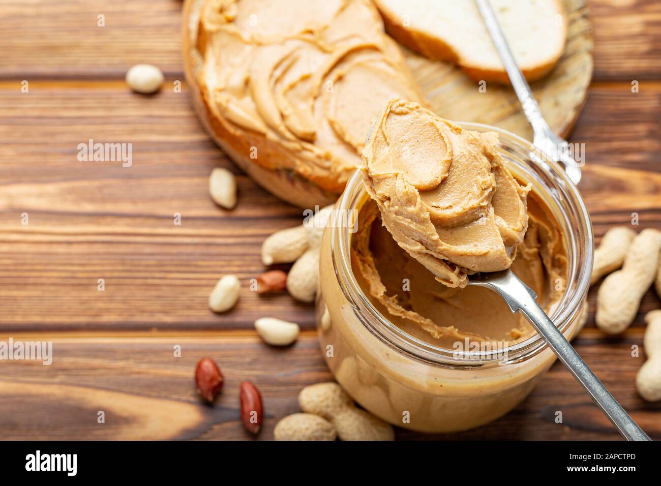 Toast con burro di arachidi. Cucchiaio e vasetto di burro di arachidi e arachidi per la prima colazione su fondo di legno marrone. Pasta di arachidi cremosa Foto Stock