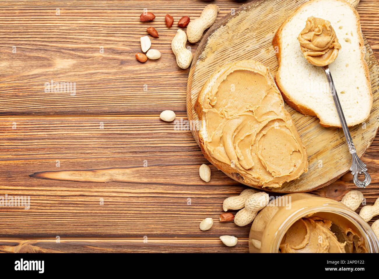 Toast con burro di arachidi. Cucchiaio e vasetto di burro di arachidi e arachidi per la prima colazione su fondo di legno marrone. Pasta di arachidi cremosa Foto Stock