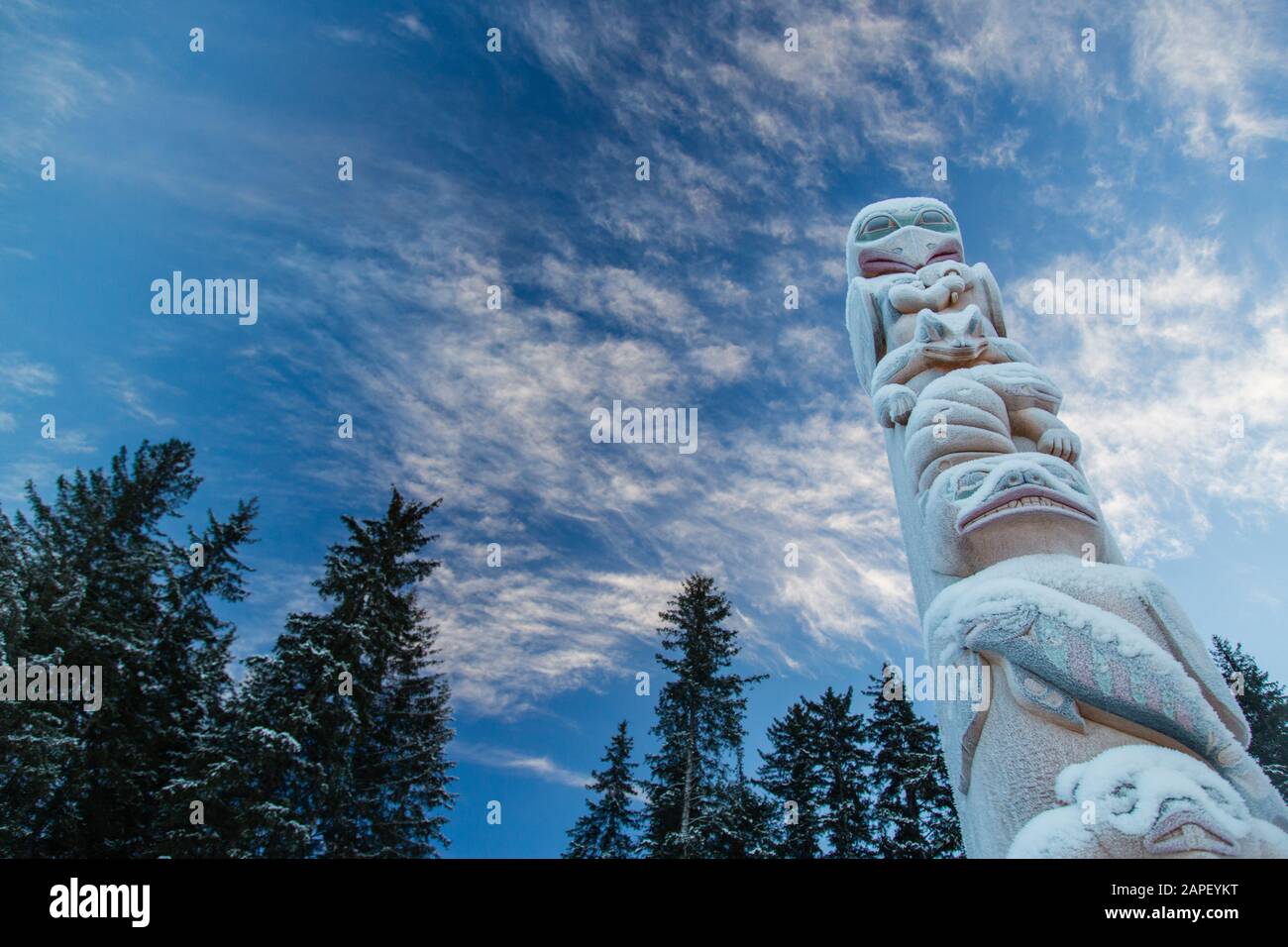 Un palo totem innevato e ricoperto di ghiaccio di fronte ad alti alberi di abete rosso e cieli blu. Foto Stock