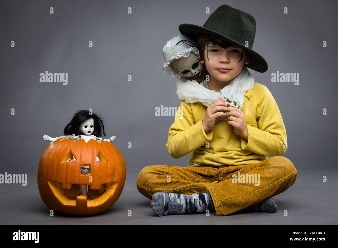 Allegro ragazzino in un cappello tiene un guazzo, zucca con bambola spaventosa accanto a lui, sfondo grigio. Festa di Halloween Foto Stock