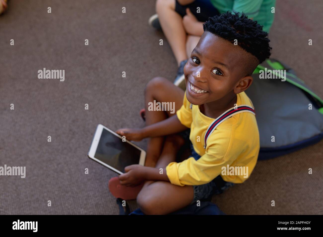 Schoolboy seduto a gambe incrociate sul pavimento utilizzando un computer tablet in una scuola elementare classroo Foto Stock